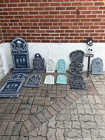 Overview of 12 various sized foam tombstones arranged against a white wall on paved ground. Includes several black, gray, white, and green tombstones with Halloween motifs.