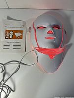 White LED beauty mask with red LED lighting illuminated, connected with cables alongside instruction booklet.