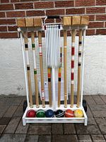 Full view of croquet set in white wooden stand showing 6 wooden mallets with colored stripes on handles, 6 colored croquet balls lined up at bottom, white metal wickets folded in center, and chrome handle on stand.