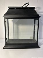 Front view of black wrought iron lantern showing glass panel, decorative latch and squared base.