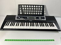 Front view of Yamaha PSR-E223 keyboard showing 61 white and black keys with attached music stand and measuring scale in front.