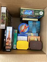 Top view of box showing multiple 1000-piece jigsaw puzzle boxes with images like the Eiffel Tower alongside Trivial Pursuit and plastic puzzle accessories in colorful containers.