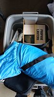 Blue sleeping bag or camping mat atop a gray plastic bin with propane lantern box partially visible inside