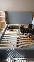 Gray double bed frame with wooden slat base partially dismantled showing storage drawers underneath and surrounding bedroom setting.