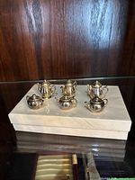 Photo shows six miniature silver tea set pieces placed on a white stand against a wood background, showcasing detail and shape of each item.