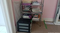 Metal chrome shelving unit holding papers, notebooks, office items with black plastic drawer unit below