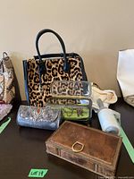 Collection showing leopard print Michael Kors handbag, silver clutch, glass jewelry box, brown leather box and partial snakeskin handbag.
