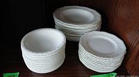 Three stacks of white ceramic dinnerware: small plates, soup bowls, and dessert bowls, all embossed with the Maria Weiss pattern around the edges.