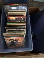 Top view of a plastic bin filled with multiple Disney music CDs in jewel cases, stacked vertically showing spine titles of various albums and soundtracks.