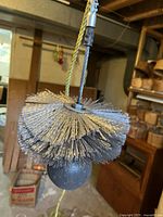 Close-up of chimney sweeping brush featuring three layers of wire bristles attached to a metal frame with a black weighted ball beneath and connected to a rope.
