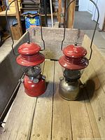 Two Coleman lanterns positioned side by side on a wooden surface, showing one with a chrome base and another fully red painted base
