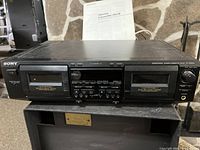 Full front view of Sony stereo cassette deck showing dual cassette slots, control panel, auto record level knob, and original manual behind unit