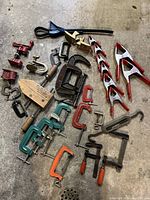 Photo showing assortment of clamps including spring clamps, various C clamps in different sizes and paint colors, one pipe clamp hook, and woodworker clamps with wooden handles.