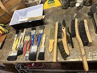 Eight paint brushes and seven wire brushes laid out on a workbench showing handle colors and bristle types