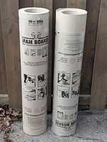 Two standing rolls of Ram Board temporary floor protection against a wooden backdrop showing product information and usage graphics.