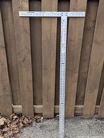 Full view of the 48-inch aluminum drywall square resting against a wooden fence showing its shape and measurement markings.