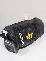 Side angle of the black leather Adidas duffel bag showing yellow trefoil logo and adidas branding on handle and side pocket.