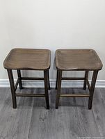Two dark brown bamboo wood bar stools with square contoured seats, standing on gray wood floor against a white wall.