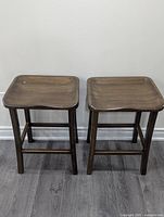 Two dark brown bamboo wood bar stools with square seats and four legs each, shown against a wall on wood flooring.