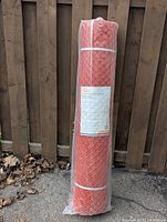 Full view of packaged orange barrier fence roll standing upright against a wooden fence, showing plastic mesh and packaging.