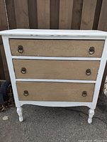 Full front view of white vintage dresser with visible chipping paint and three drawers with ornate front panels and metal ring handles.