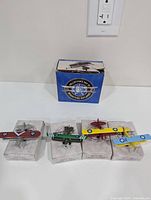 Overview of all four miniature biplane models with original blue collector's box in background. Planes mounted on clear plastic display bases with printed cardboard beneath. Box with some wear.