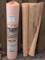 Two upright rolls against a wooden fence: sealed Schluter-DITRA5M roll on left and open Schluter-KERDI membrane roll on right