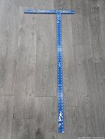 Blue metal T-shaped drywall square with white measurement markings, showing signs of use and paint chipping, lying on a wooden floor.