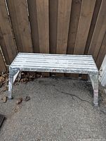Single foldable metal bench with visible signs of use on an outdoor pavement area with wooden fence background.