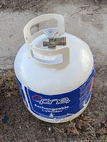 Top-front view of a white, preowned, empty propane tank with blue label and a metal valve on top, sitting on ground outdoors.