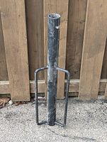 Full view of black metal fence post driver standing vertically showing tubular body with side handles against wooden fence background.