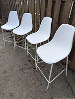 Four white plastic/resin bar height chairs with white metal legs set outdoors showing front and side views on asphalt near a brown wooden fence.