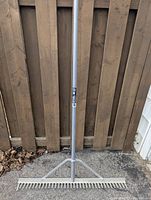 Full view of the 36-inch aluminum landscaping rake leaning against a wooden fence, showing the long metallic handle and the wide rake head with multiple tines.