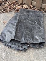 Folded large black tarp laid on the ground showing metal ring tie downs and creased, worn surface.