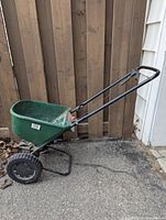Scotts Basic Lawn Seeder with green hopper and black metal handles and wheels shown on driveway beside wooden fence and wall.