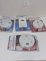 Four Kidde detectors sealed in retail packaging placed on a white surface. Includes combo smoke and CO detectors and specialized gas detector.