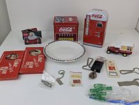 Photo showing wide arrangement of Coca Cola collectibles including tins, plate, Coke van model, miniature bottles, bottle openers and pin.