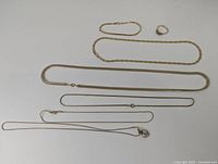 Seven gold-toned chains, a ring, and a bracelet displayed on white surface. Various chain styles and thicknesses, pendant with clear stones visible.