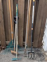 Photo showing six different landscaping and gardening tools standing against a wooden fence, including leaf rake, garden rakes, cultivators, and hoe.