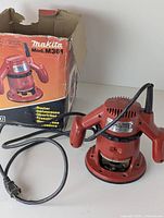 Makita M361 wood router next to its original box showing front and side views of router and box labeling.