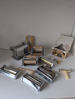 Wide view of the pasta making tools showing several roller machines, cutter parts, a ravioli mold with wooden handles, and a plastic dough cutter.