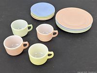 Set of 3 miniature teacups, 1 creamer, 4 saucers, and stack of 4 plates all in pastel colors on a dark surface.