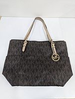 Front view of the brown Michael Kors tote bag showing full MK logo pattern, beige leather handles with adjustable buckles, and round MK logo charm hanging from one handle.