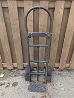 Photo of a preowned moving dolly with a black metal frame positioned upright against a wooden fence outdoors. Shows two dry rubber tires, rust on the metal parts, and a missing pin or two.