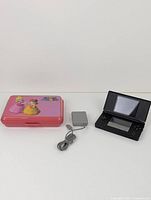 Front view of Nintendo DS console, power adapter, and pink Princess Peach carrying case displayed side by side on a white surface.