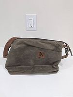 Full view of olive green shoulder bag lying flat showing front, brown adjustable strap, and leather patch logo.