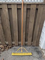 Push broom standing upright against a wooden fence with a detailed view of the handle and yellow bristles.