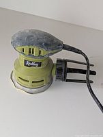 Green and black Radley electric palm sander with round sanding base and power cord, showing top and side view.