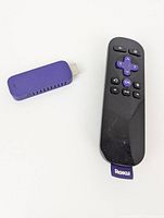 Photo shows a purple Roku USB streaming device alongside a black Roku remote control on white background.