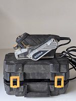 Side view of RONA palm planer tool placed on black plastic carrying case with yellow latches, showing overall tool shape and color.